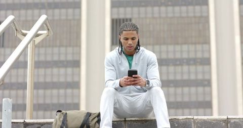 African American Man Sitting on Urban Ledge Checking Phone in Gray Tracksuit with Smartwatch