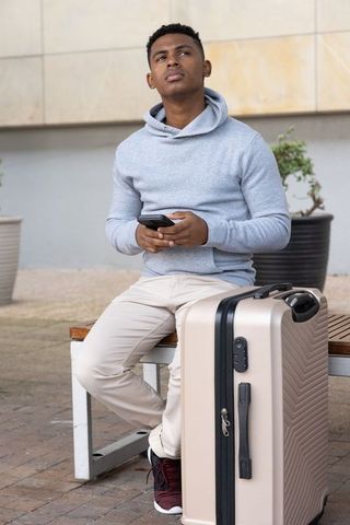 African american traveler waiting on bench with carry-on suitcase and smartphone