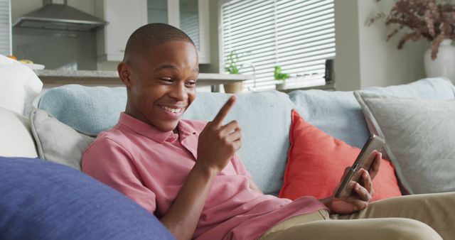 Young man in a casual pink shirt smiling and pointing while holding a smartphone during a video chat at home. Ideal for illustrating concepts of modern communication, technology usage, social media interaction, and casual, relaxed home settings.