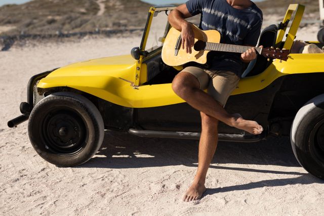 Low section of caucasian man sitting on beach buggy playing guitar on sunny beach. beach stop off on summer holiday road trip.