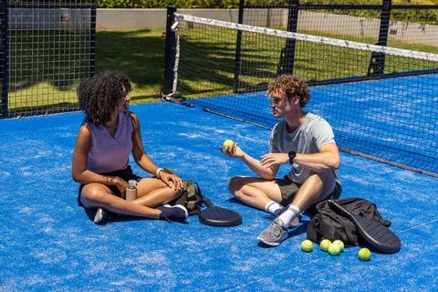 Diverse friends sitting on blue padel court chatting and holding ball and paddles