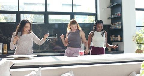 Diverse women dancing at kitchen island celebrating with drinks in bright modern home
