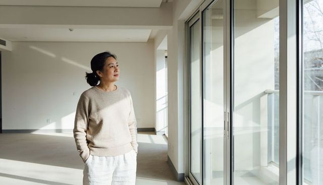 Standing asian woman gazing through sunlit apartment windows embracing calm minimal interior