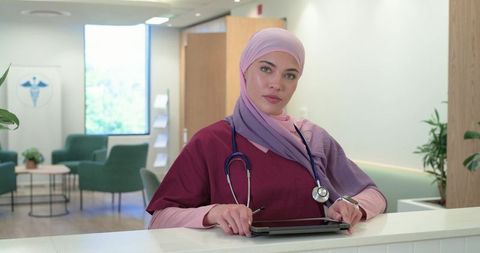 Muslim female nurse in hijab using tablet at modern clinic reception desk wearing scrubs