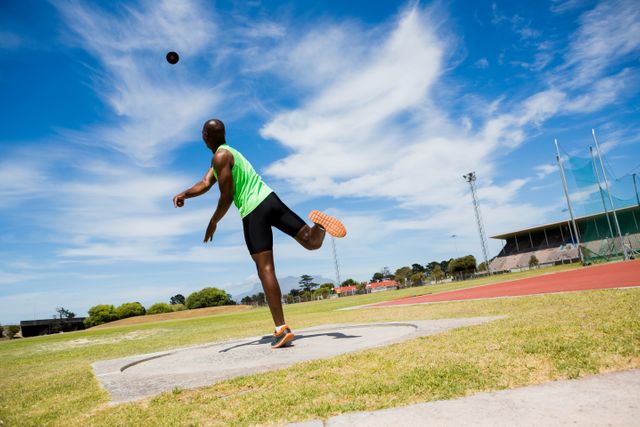 Male athlete in green tank top and black shorts throwing shot put ball in stadium. Clear blue sky with scattered clouds in background. Ideal for use in sports-related content, athletic training materials, fitness promotions, and competitive event advertisements.