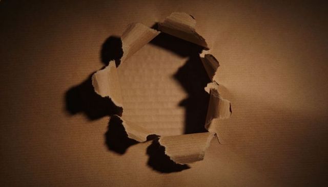 Revealing ragged circular tear in corrugated cardboard with curled flaps and warm shadows