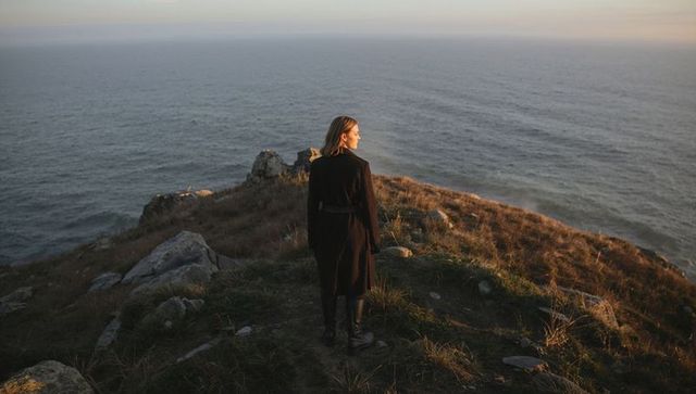 Solitary woman standing on coastal cliff facing ocean horizon at golden hour