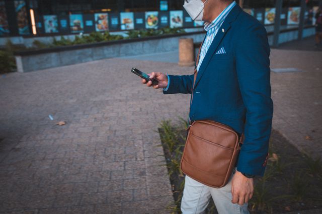 Businessman in blue blazer and white pants walking outdoors in a corporate park while using a smartphone. He is wearing a face mask, indicating adherence to COVID-19 safety measures. This image can be used to illustrate topics related to business, technology, pandemic safety, urban lifestyle, and professional settings.