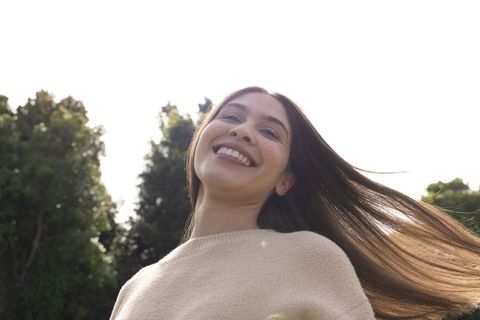 Asian woman smiling and looking upward in sunlit park with flowing hair and beige sweater