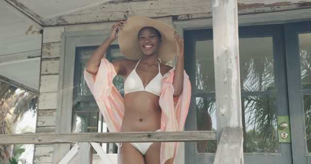 Smiling woman wearing a white bikini and colorful cover-up stands on a beachfront balcony, adjusting her straw hat and enjoying a sunny day. Ideal for promoting summer vacations, beachwear fashion, tropical getaways, or positive lifestyle themes.