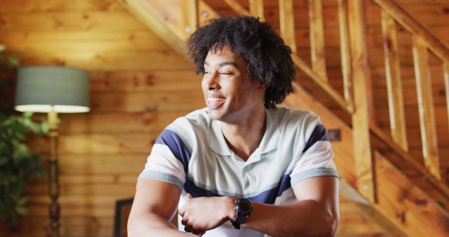 Smiling man sits comfortably in a rustic wooden cabin, displaying a sense of relaxation and happiness. Ideal for promoting leisure activities, vacation rentals, or home comfort products. Can be used in lifestyle blogs or advertisements highlighting serene, cozy living spaces.