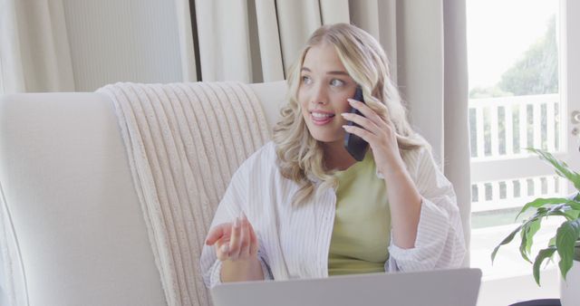 Young woman with long blonde hair speaking on mobile phone while sitting in a white armchair at home. Dressed casually in a light green shirt and white over-shirt, typing on laptop. Ideal for conveying themes of remote work, technology in everyday life, modern communication methods, or relaxed domestic settings.