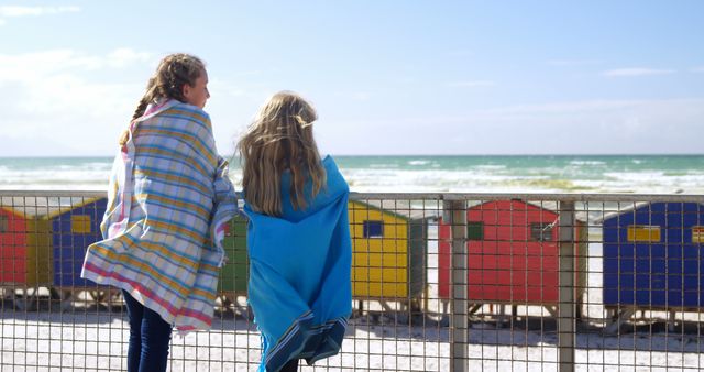 Back view of caucasian girls standing wrapped blankets and looking on sea on sunny promenade. Childhood, friendship, free time, summer, travel, vacations and lifestyle, unaltered.