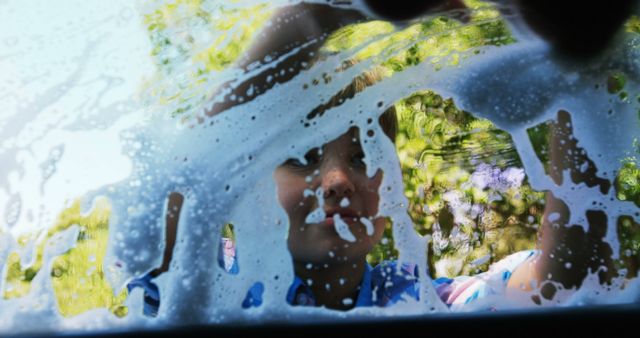 A young child is seen through a car window covered in soapy water, helping with a car wash. The playful interaction captures a moment of childhood joy and participation in a common household chore.