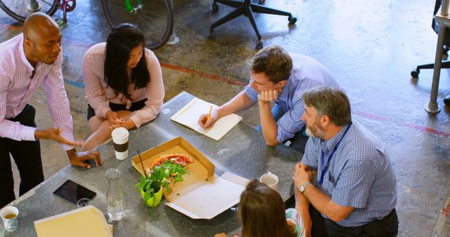 Business professionals from diverse backgrounds gather around a table with pizza for an interactive and engaging team meeting in an open workspace. They are brainstorming and collaborating, emphasizing a casual and productive work culture. Perfect for illustrating modern office environments, teamwork, and business strategy concepts.