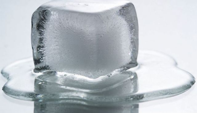 Melting single clear ice cube on reflective surface with puddle and trapped air bubbles