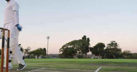 Diverse male cricketers walking by stumps wearing white kit at sunset on grass pitch