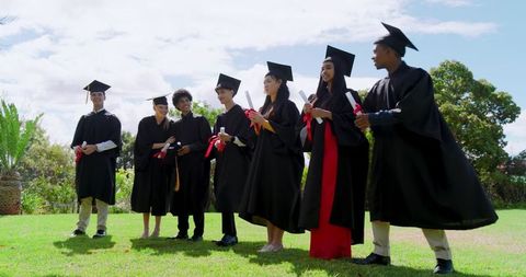 Diverse graduates celebrating outdoors wearing graduation gowns and caps holding diplomas