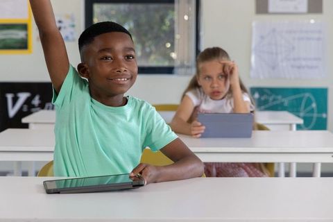 Black boy raising hand using tablet in classroom, white girl studying on tablet
