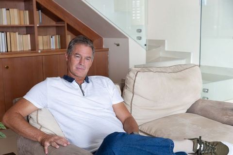 Mature man relaxing on beige sectional sofa in modern living room with bookshelf and stairs