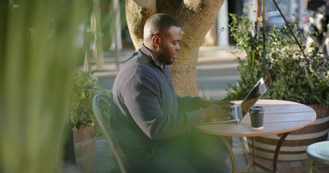African American Man Working Remotely on Laptop at Urban Cafe Patio Surrounded by Greenery