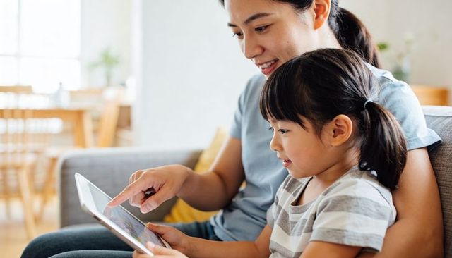 Smiling asian mother guiding daughter on tablet learning together on gray couch at home
