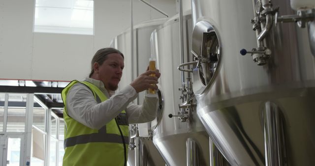 Brewery worker wearing safety vest inspecting beer quality in industrial brewery. Ideal for use in articles about beer production, brewing industry, quality control processes, and industrial workplace environments.