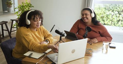 Multiracial female hosts recording casual home podcast at dining table with laptop