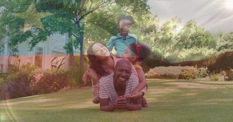 Laughing family stacking together on sunlit lawn, parents and children playing outdoors