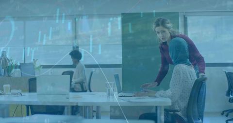 Collaborating colleagues wearing hijab reviewing laptop with data overlay in modern office
