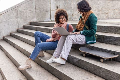 Friends collaborating on laptop on urban steps with skateboard and autumn scarves