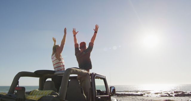 Happy caucasian gay male couple standing in car raising arms and holding hands on sunny day at beach. summer road trip and holiday in nature.