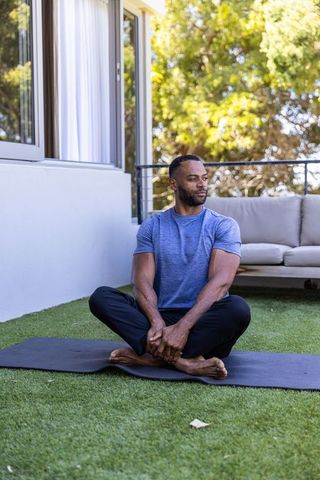 African american man practicing seated yoga on black mat on sunlit patio with lounge