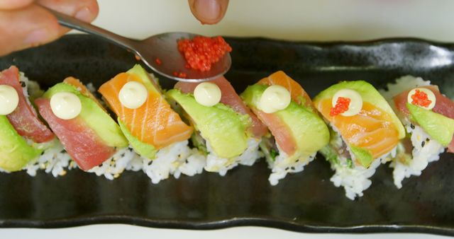 A spoon garnishes sushi with roe, adding a flavorful touch. The sushi arrangement showcases a fusion of colors and textures on a sleek plate.