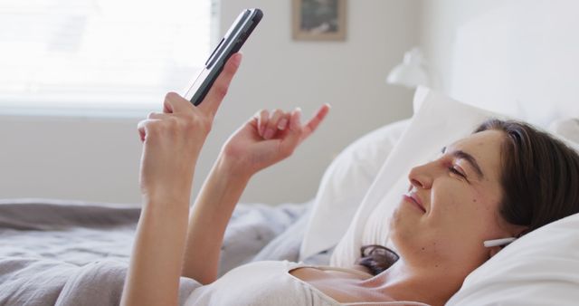 Happy caucasian woman relaxing, lying in bed and using smartphone. Spending quality time at home concept.