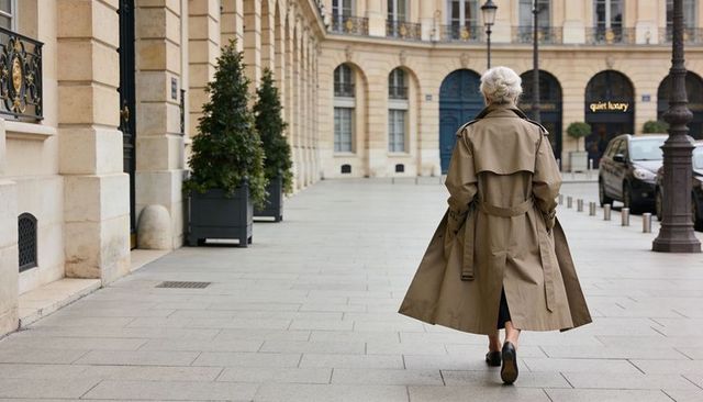 Senior woman walking in tan trench coat along elegant arcaded city street, rear view