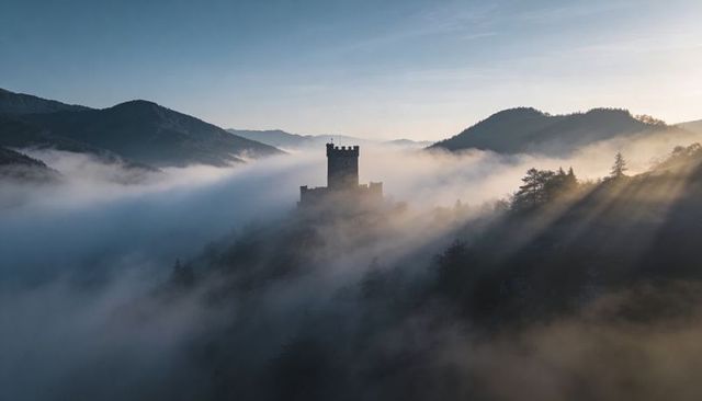 Medieval tower emerging from morning fog on pine ridge with sunrays over valley