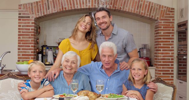 A joyful multi-generational family gathers around a dinner table in a cozy kitchen. Grandparents sit in the foreground with their young grandchildren, while the parents stand behind, all smiling. Ideal for themes of family togetherness, bonding, intergenerational relationships, and celebratory moments at home.