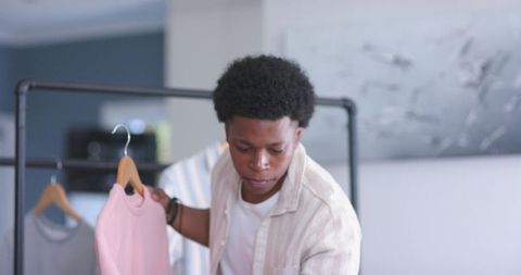 Young male stylist inspecting pink top on wooden hanger while arranging boutique rack