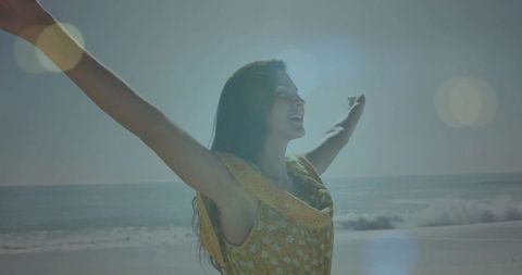 Woman stretching arms facing sun on beach in yellow floral dress, joyful backlit moment