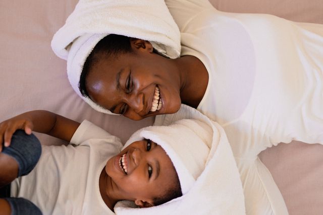 Mother and daughter lying on bed, both wearing white turbans, sharing a joyful moment. Ideal for use in family-oriented content, parenting blogs, advertisements promoting family products, or articles about family bonding and home life.