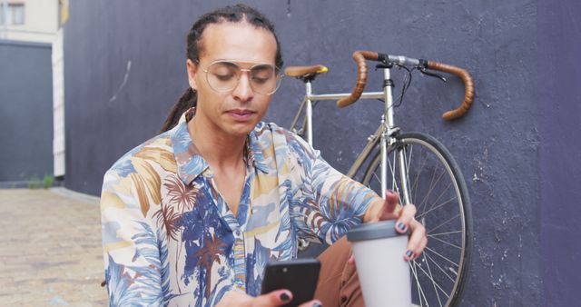 Front view close up of a biracial man with long dreadlocks out and about in the city on a sunny day, sitting in the street, drinking coffee, using a smartphone, with his bicycle leaning against the wall next to him in slow motion.