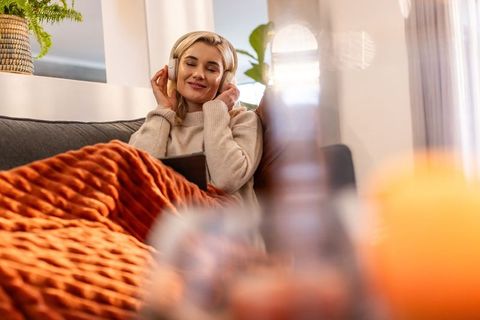 Relaxing woman in thirties listening to music with headphones on cozy rust blanket sofa