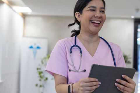 Smiling female nurse in pink scrubs holding tablet in modern clinic with stethoscope