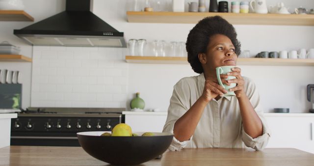 African American woman holding coffee cup while sitting in modern kitchen. Background includes a stove with hood, white tiled backsplash, and open shelves with cups and bowls. Useful for lifestyle, home, and morning routine concepts.