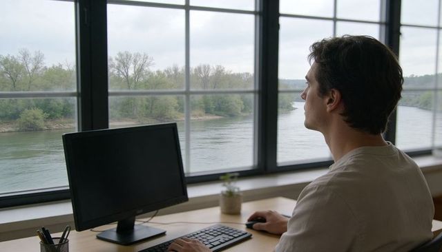 Young man gazing at river from minimalist riverside workspace with desktop monitor and plant