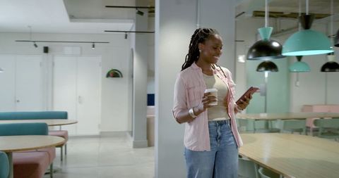 Smiling African American woman holding coffee and checking smartphone in modern coworking lounge