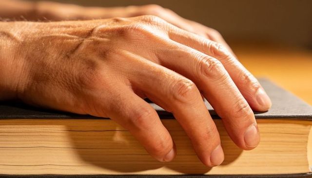 Adult hand resting on closed hardcover book with yellowed pages on warm wooden desk