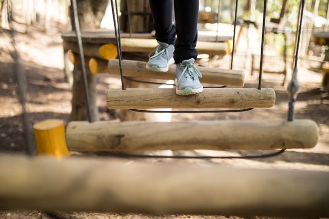 Child crossing a wooden zip line bridge in an adventure park, focusing on the feet and legs. Ideal for use in articles or advertisements about outdoor activities, children's recreation, adventure parks, and promoting physical fitness and balance skills in kids.