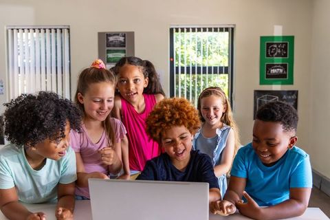 Diverse children collaborating around laptop in bright classroom enjoying digital learning